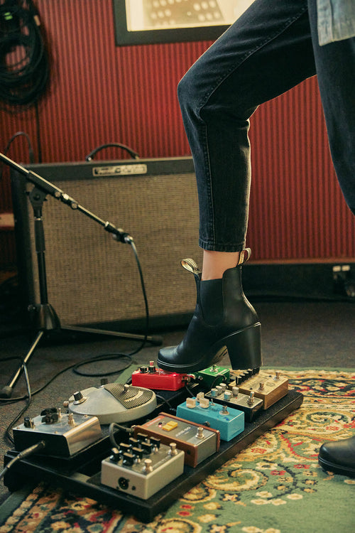 A person wearing black heeled boots steps on a pedalboard with various colorful guitar effects pedals. An amplifier and cables are visible in the background, placed on a patterned rug.