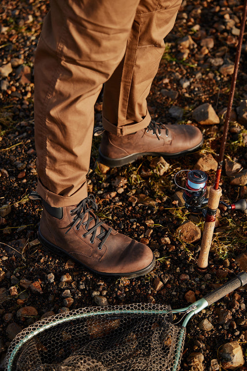 A person wearing brown pants and brown boots stands on rocky ground beside a fishing rod and a net, with some moss and small stones scattered around.