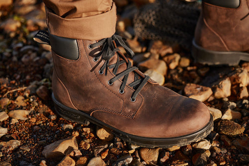 A close-up of a person wearing brown leather hiking boots and tan pants, standing on a rocky ground. The boots have black laces and a black padded collar.