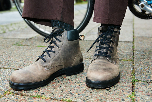 A person wearing dark brown pants and gray lace-up boots stands on a paved sidewalk next to a bicycle wheel.