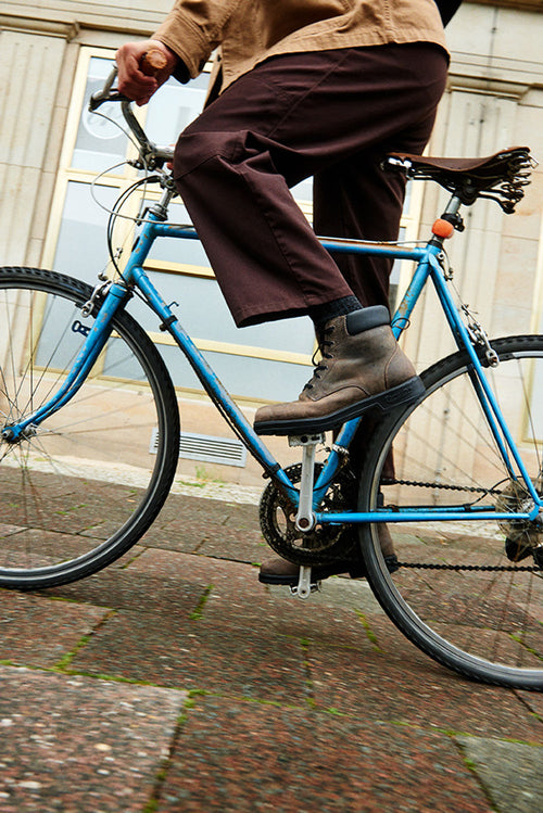A person wearing brown pants and boots is riding a blue bicycle on a city sidewalk near a building with large windows. The photo is taken from a low angle, focusing on the bike and the riderâ€™s legs.
