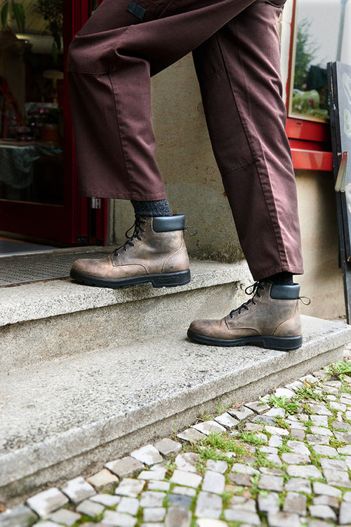 A person wearing brown pants and brown lace-up boots is stepping up a set of stone stairs outside a building, with cobblestone pavement visible at the bottom.