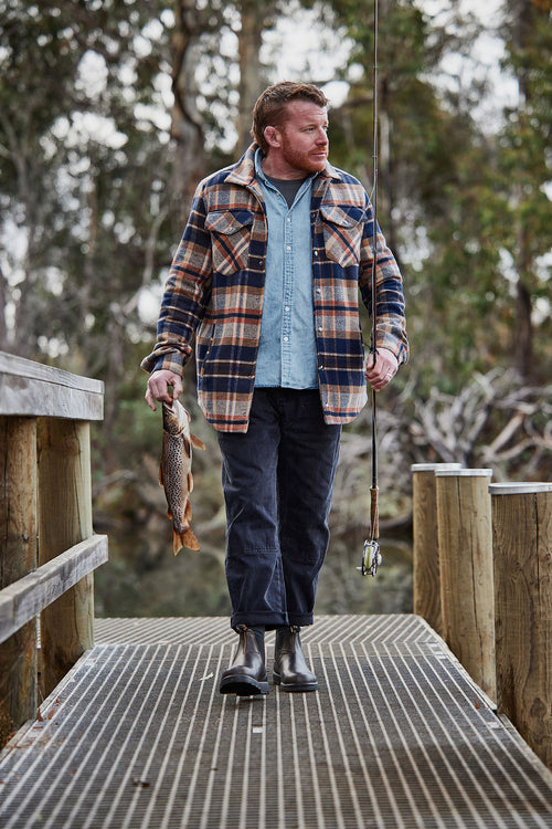 A man in a plaid jacket and boots stands on a dock holding a fishing rod in one hand and a fish in the other, with trees and water in the background.