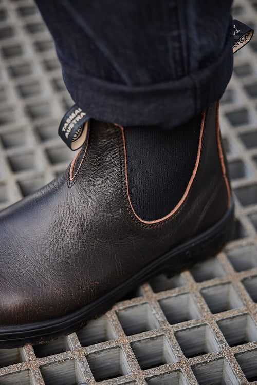 A close-up of a persons foot wearing a brown leather Chelsea boot with black elastic side panels and dark jeans, standing on a metal grid surface.