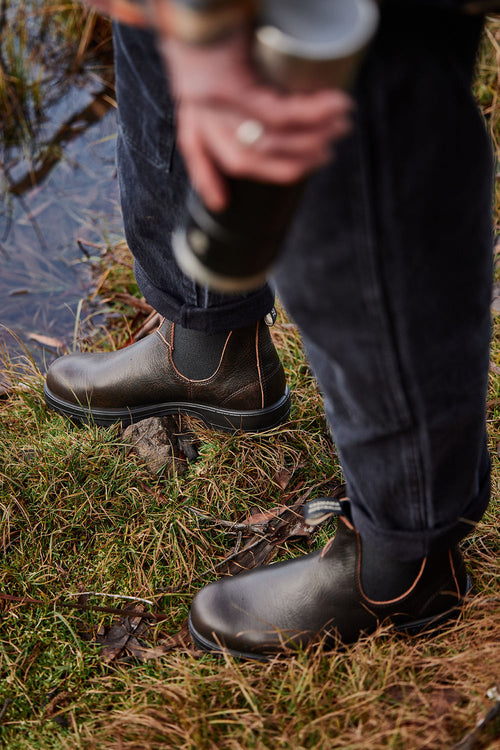 Person wearing brown leather boots stands on grassy ground near water, holding a metallic thermos. Only the lower half of the person is visible.
