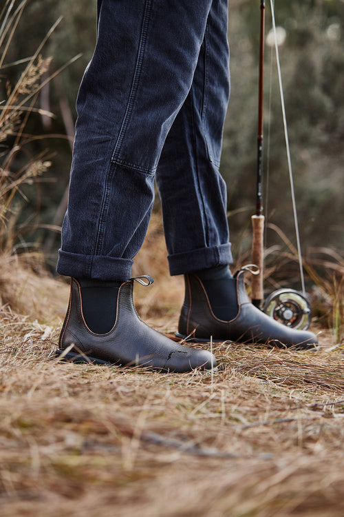 A person standing on dry grass wearing dark jeans and brown leather boots, holding a fishing rod. The setting appears to be outdoors, possibly near a river or lake.