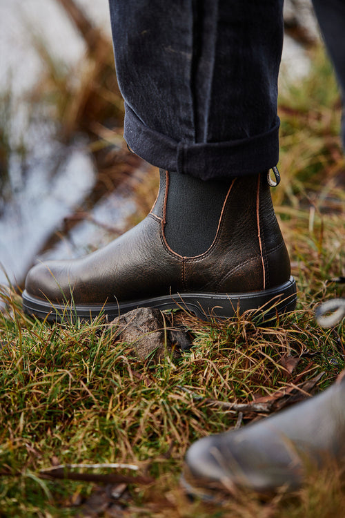 A close-up of a person wearing dark brown leather Chelsea boots and cuffed black jeans, standing on grass near water.