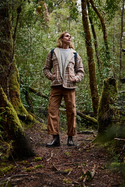 A person wearing a plaid jacket, brown pants, and boots stands on a forest trail, looking upward with hands in pockets, surrounded by tall trees and lush green foliage.
