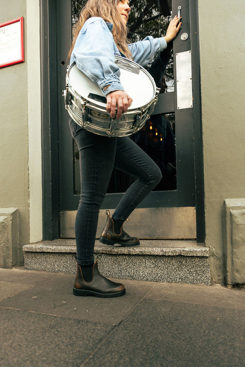 A person in a denim jacket and black jeans holds a snare drum while opening a black door and stepping up onto a small stoop.