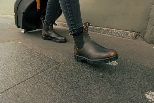 A person wearing dark brown Chelsea boots and black jeans walks on a sidewalk, pulling a black wheeled suitcase behind them.