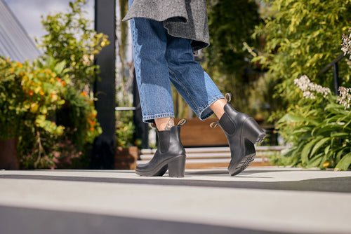 A person wearing cropped blue jeans, a gray coat, and black heeled ankle boots walks outdoors on a sunny day, with greenery and plants in the background.