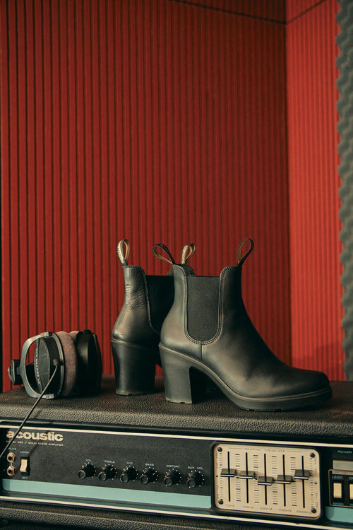 A pair of black heeled Chelsea boots and black headphones rest on top of an acoustic amplifier in a music studio with red and black soundproofed walls.
