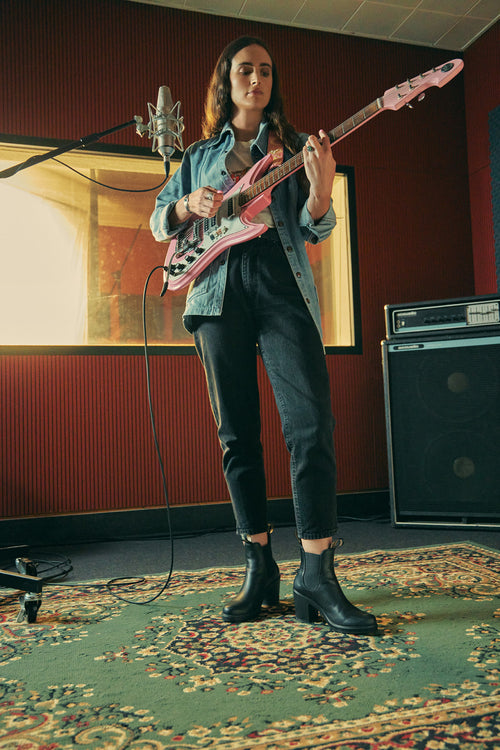 A woman stands in a music studio playing a pink electric guitar, wearing black jeans, a denim jacket, and black boots. A microphone is positioned in front of her, and amplifiers are visible in the background.