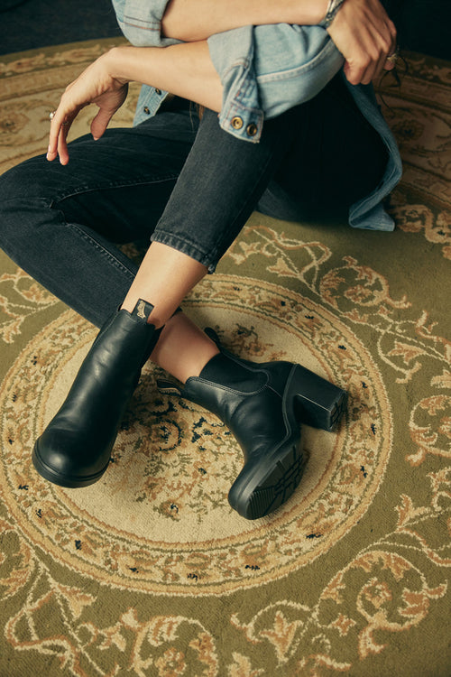 A person sitting cross-legged on an ornate green rug, wearing black heeled ankle boots, black jeans, and a light denim jacket with sleeves rolled up. A small tattoo is visible above one boot.