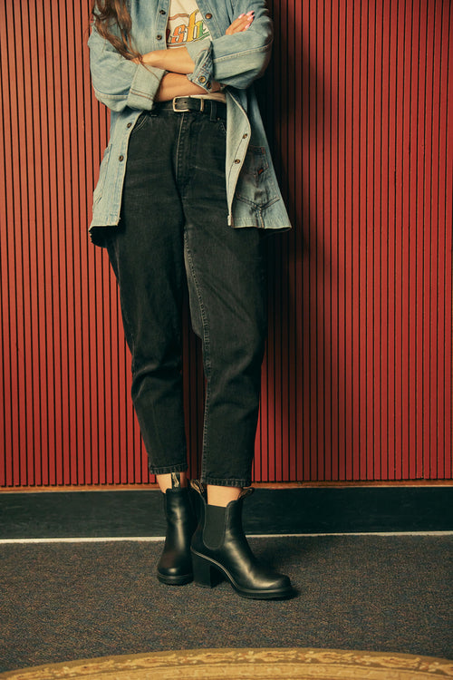A person stands against a red, vertically-striped wall with arms crossed, wearing a denim jacket, black high-waisted jeans, and black ankle boots. Their face is not visible in the image.