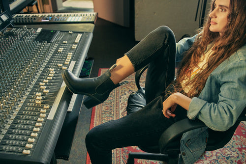 A woman with long hair sits in a music studio, resting one booted foot on the mixing console. She wears jeans and a denim jacket, looking thoughtful in a room with audio equipment and a patterned rug.