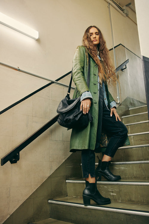 A woman with long wavy hair stands on a stairway, wearing a green leather coat, black boots, and carrying a large black bag. She looks confidently at the camera, with one foot on a higher step.