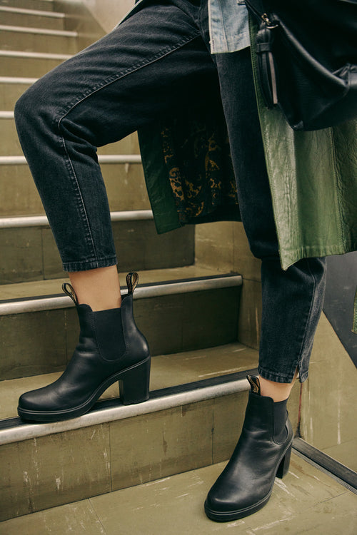 Person wearing black heeled ankle boots and dark rolled-up jeans, standing on stairs; part of a green coat and a black bag are visible.