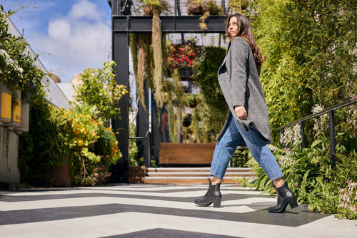 A woman in a gray coat, blue jeans, and black boots walks confidently along a modern garden pathway with greenery and hanging plants, under a partly cloudy sky.
