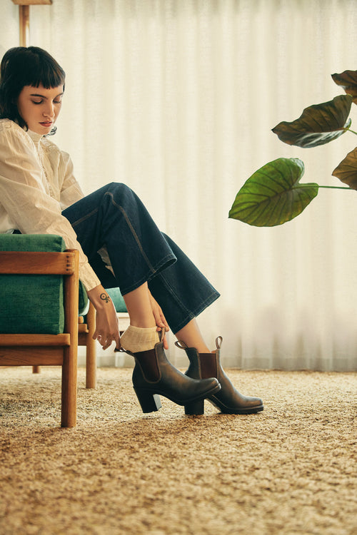 A woman with short dark hair sits on a green sofa in a sunlit room, putting on dark heeled ankle boots. She wears a white blouse, rolled-up jeans, light socks, and is framed by a large leafy plant and sheer curtains.