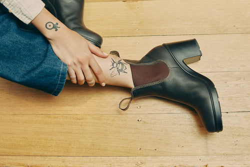 A person sitting on a wooden floor, wearing blue jeans and black heeled ankle boots, with a bee tattoo on their ankle and another tattoo on their hand.