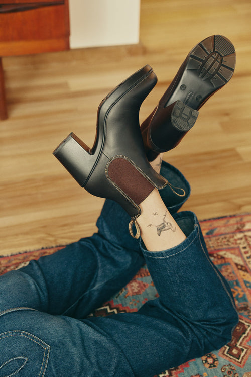 A person in blue jeans lies on a patterned rug, feet raised, showing black heeled boots and an ankle tattoo of a kangaroo. The wooden floor and part of a cabinet are visible in the background.