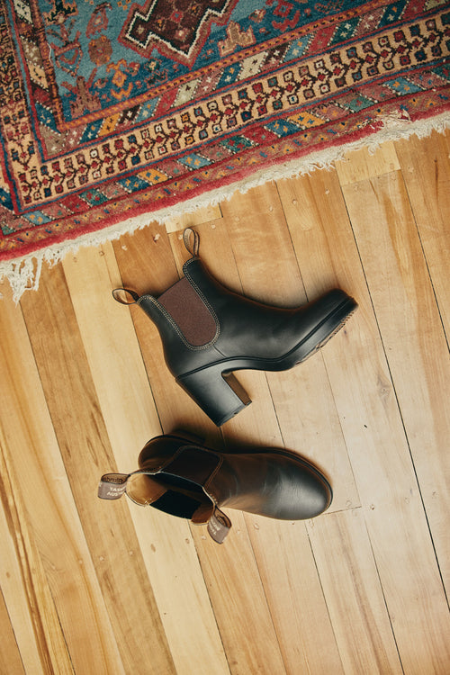 A pair of black heeled Chelsea boots rest on a light wooden floor near the corner of a colorful, patterned rug with fringed edges.