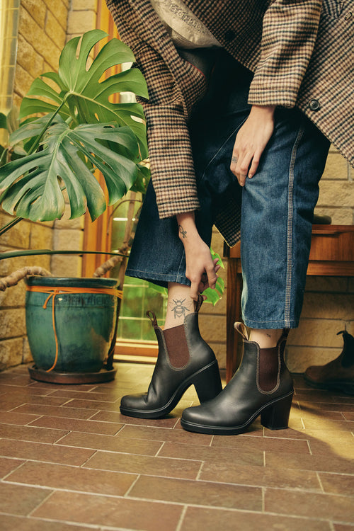 A person wearing rolled-up jeans, a plaid coat, and black heeled boots stands indoors next to a potted plant, adjusting one boot. A tattoo is visible on their ankle.