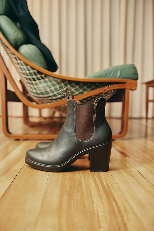 A black high-heeled Chelsea boot with brown elastic side panels sits on a light wooden floor, with a cushioned lounge chair in the background.