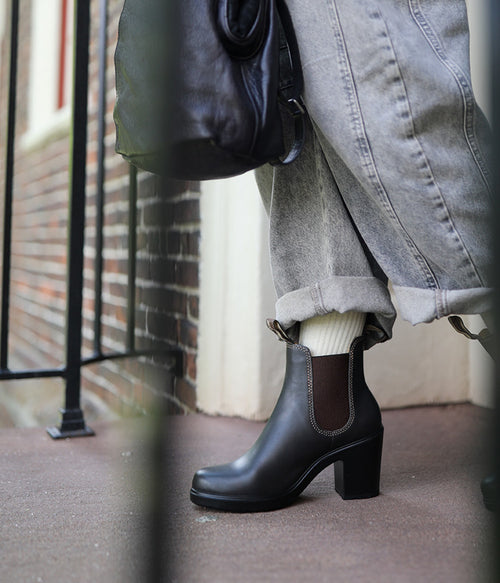 A person wearing light gray cuffed jeans, white socks, and black heeled ankle boots stands on a city sidewalk, holding a black bag. The image is framed by a metal railing and shows a brick wall in the background.