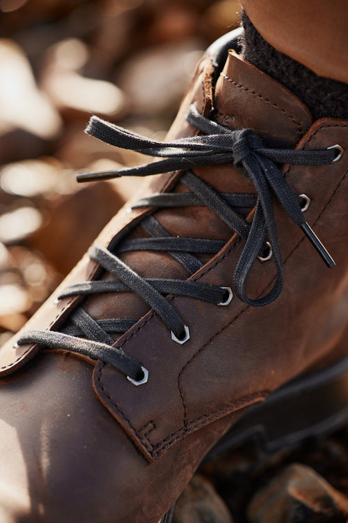 Close-up of a brown leather hiking boot with black laces, tied neatly. The boot is worn and resting on rocky ground, suggesting an outdoor setting.