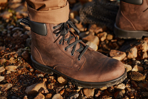 A close-up of a person wearing brown leather hiking boots and tan pants, standing on a rocky ground. The boots have black laces and a black padded collar.