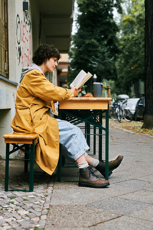 A person in a yellow coat sits at an outdoor table on a city sidewalk, reading a book. Thereâ€™s a drink and pastry on the table, and bicycles are parked in the background among trees and buildings.