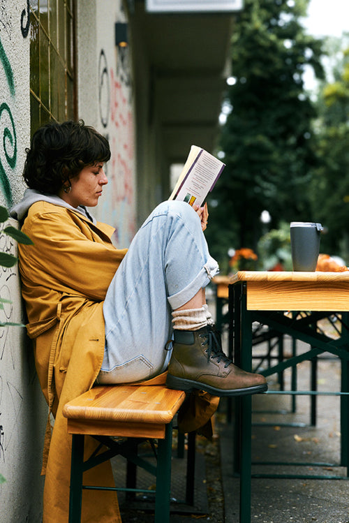 A person in a yellow coat and rolled-up jeans sits sideways on a wooden bench outside, reading a book with their knees up and a mug on the table nearby. Trees and greenery are visible in the background.