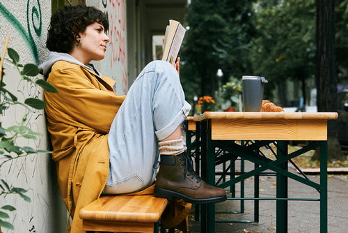 A person in a yellow coat sits on a bench outdoors, reading a book with knees drawn up. A coffee cup and croissant are on the wooden table nearby. Trees and buildings are visible in the background.