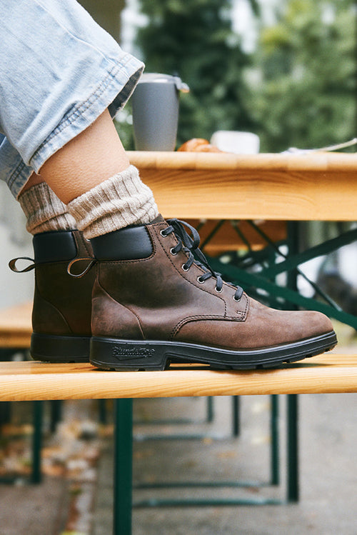 A person wearing light blue jeans, beige socks, and brown lace-up boots sits on a wooden outdoor bench. The scene is casual, with blurred greenery in the background.