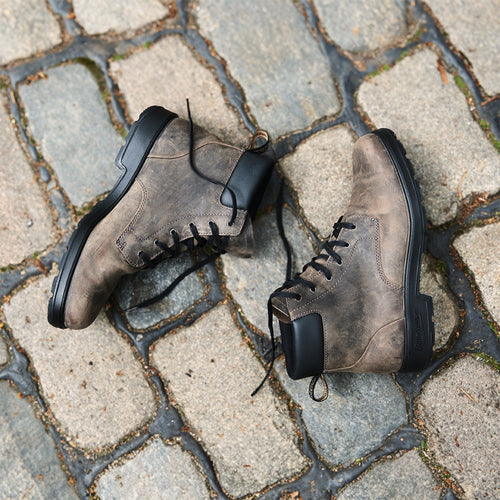 A pair of rugged brown leather lace-up boots with black soles and heels are placed side by side on a cobblestone street.