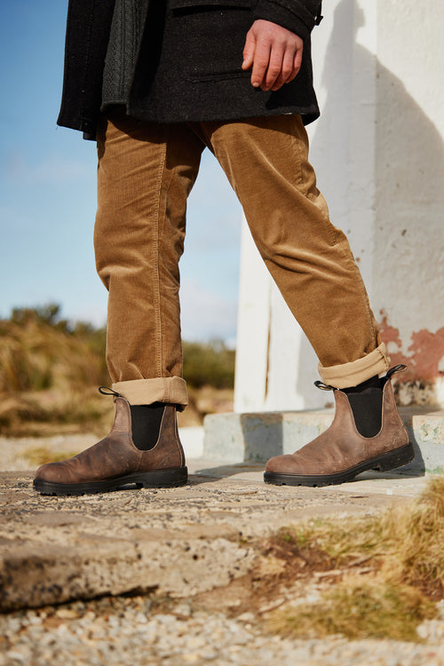 A person wearing brown Chelsea boots, tan corduroy pants, and a black coat walks outdoors on a stone path, with dry grass and a white structure in the background.