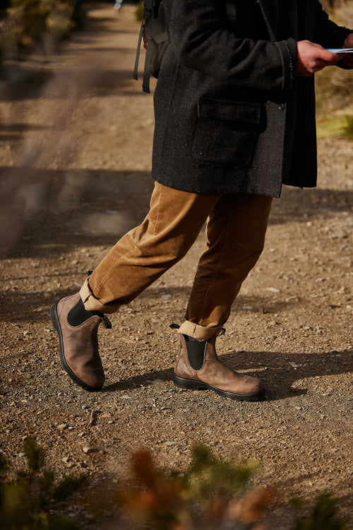 A person wearing rolled-up brown pants, brown Chelsea boots, and a dark coat walks on a dirt path, holding an object in one hand. The upper part of the personâ€™s body is not visible.