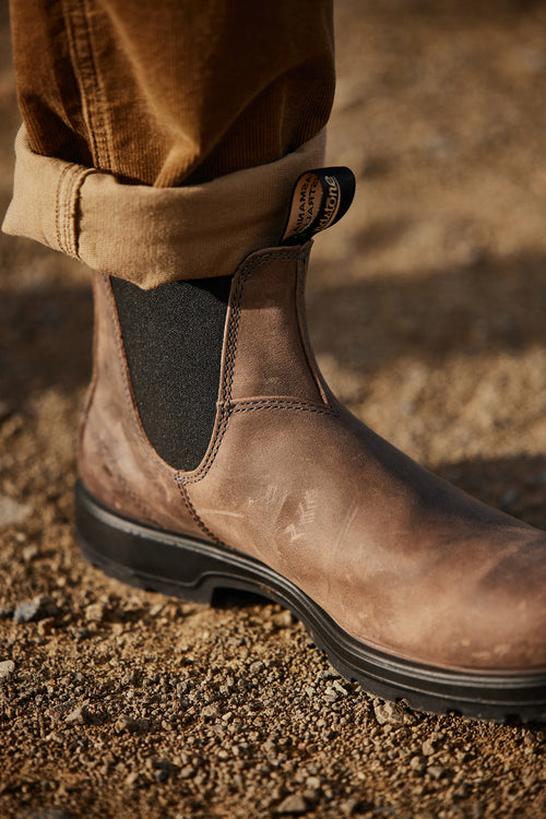 A close-up of a person wearing a brown leather Chelsea boot with black elastic side panels, standing on a gravel surface. The personâ€™s tan pants are rolled up above the ankle.