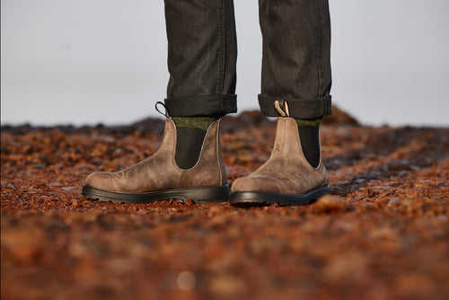A person wearing brown leather Chelsea boots and dark rolled-up jeans stands on rocky, reddish-brown ground.