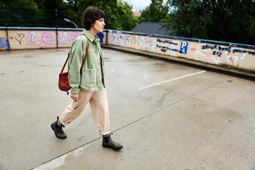 A person with short dark hair walks through an empty, outdoor parking lot, wearing a light green jacket, beige pants, dark boots, and carrying a brown shoulder bag; graffiti covers the low wall behind them.