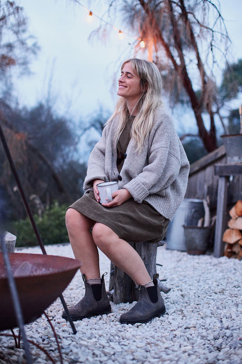 A woman with long blonde hair sits on a wooden stump outdoors, smiling and holding a mug. She is dressed in a cozy sweater, dress, and boots, with string lights and a fire pit visible in the rustic background.