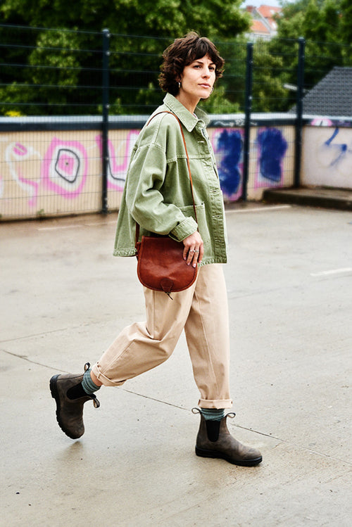 A person with short dark hair walks outdoors on a wet surface, wearing a light green jacket, beige pants, gray boots, and carrying a small brown shoulder bag. There is a graffiti-covered wall and trees in the background.