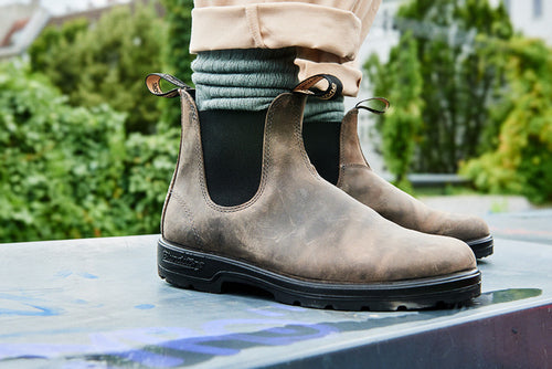 Close-up of a person wearing light brown leather Chelsea boots, green socks, and beige pants, standing outdoors on a metal surface with greenery in the blurred background.