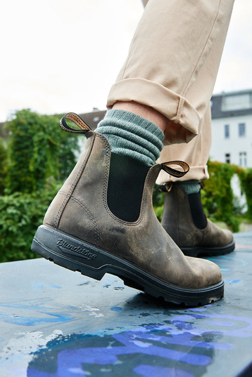 A close-up of a person wearing light brown leather Chelsea boots, green striped socks, and beige pants, standing outdoors on a painted surface with greenery and buildings in the background.