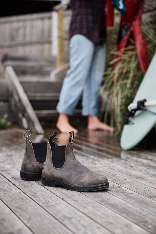 A pair of worn brown boots sits on a wooden deck. In the background, a barefoot person in rolled-up jeans stands near a green surfboard, with stairs and plants visible nearby.