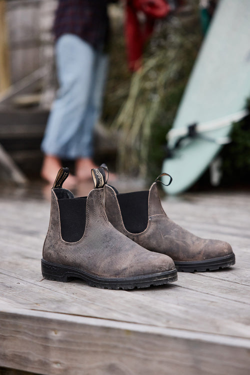 A pair of worn brown Chelsea boots with black elastic sides sits on a weathered wooden deck. In the background, a person in rolled-up jeans and a blurry surfboard are partially visible.