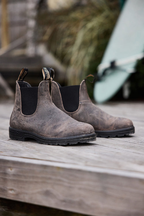 A pair of worn brown Chelsea boots with black elastic sides sits on a weathered wooden deck, with a blurred green surfboard in the background.