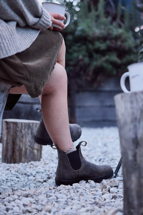 A person wearing brown boots and a skirt sits outdoors on a wooden bench, holding a mug. Another mug sits on a tree stump nearby. The ground is covered in white gravel, and green bushes are in the background.